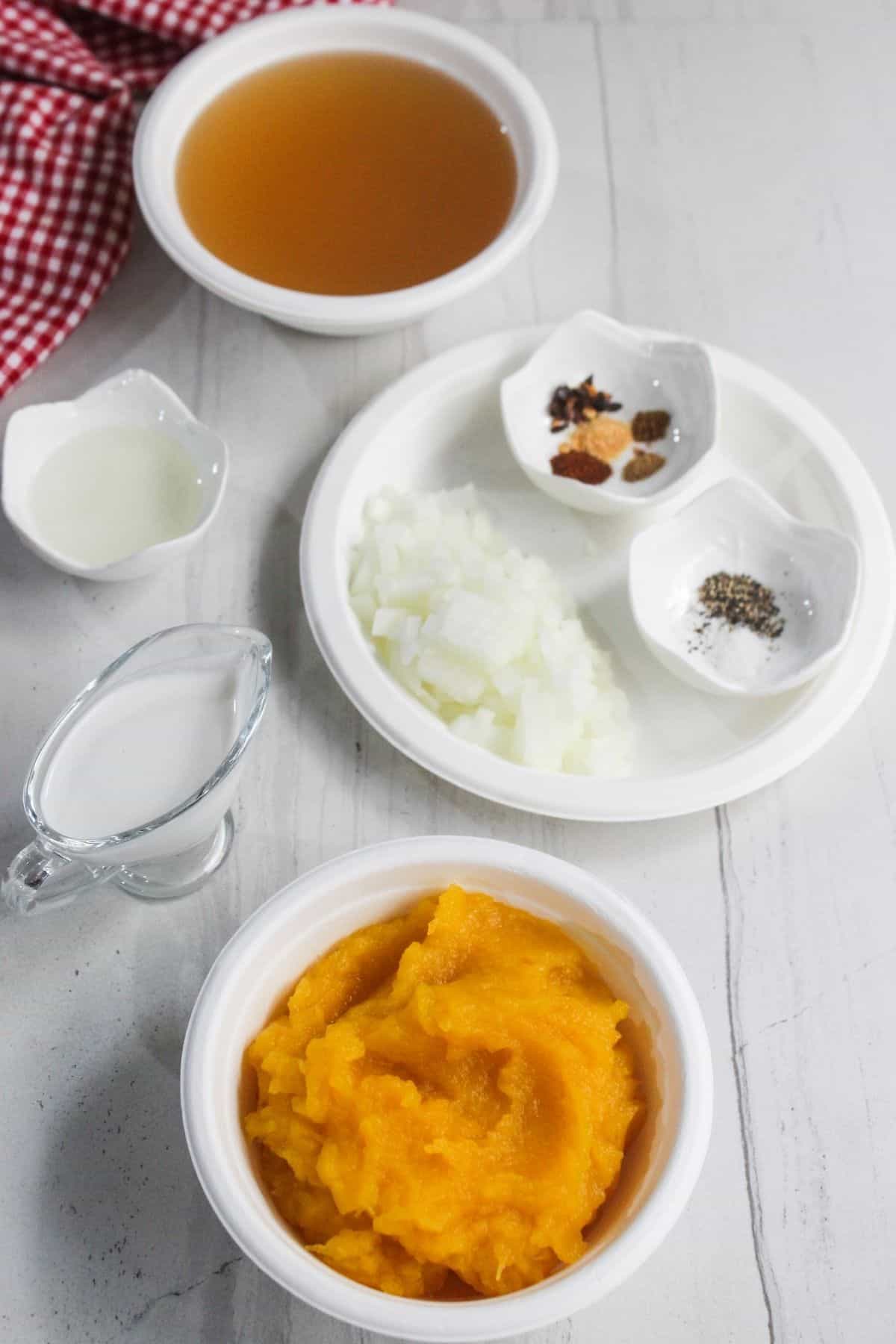 Small bowls containing pumpkin puree, chopped onions, spices, broth, cream, and vinegar are arranged on a white tabletop.