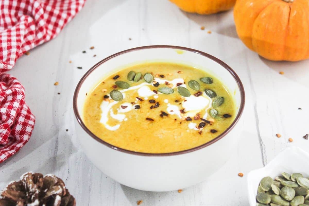 A bowl of pumpkin soup topped with cream, pumpkin seeds, and red pepper flakes, placed on a white surface with small pumpkins and a red checked cloth nearby.