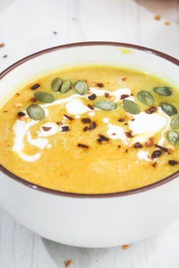 A bowl of creamy pumpkin soup garnished with seeds and a swirl of cream, placed on a white surface with a red cloth and pumpkin nearby.