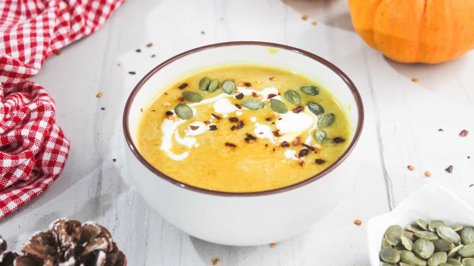 A bowl of creamy pumpkin soup garnished with seeds and a swirl of cream, placed on a white surface with a red cloth and pumpkin nearby.
