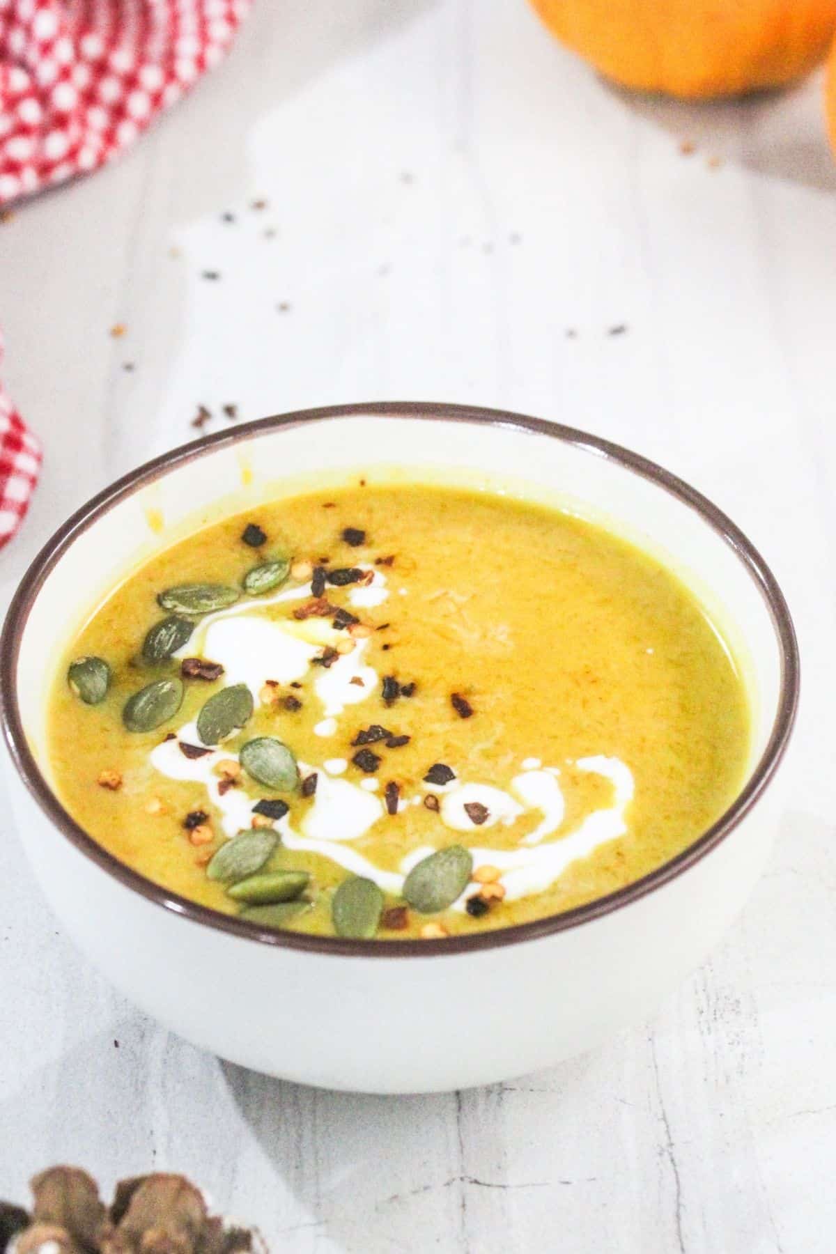 A bowl of creamy orange soup garnished with a swirl of cream, pumpkin seeds, and crushed red pepper flakes on a white surface.