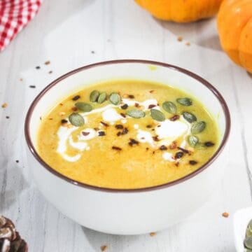 A bowl of pumpkin soup topped with cream, pumpkin seeds, and spices sits on a white table.
