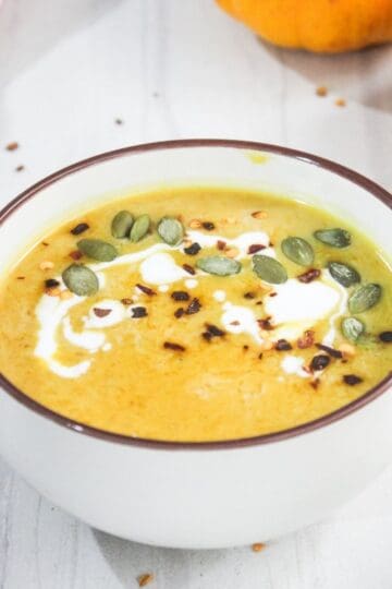 A bowl of pumpkin soup topped with cream, pumpkin seeds, and spices sits on a white table.