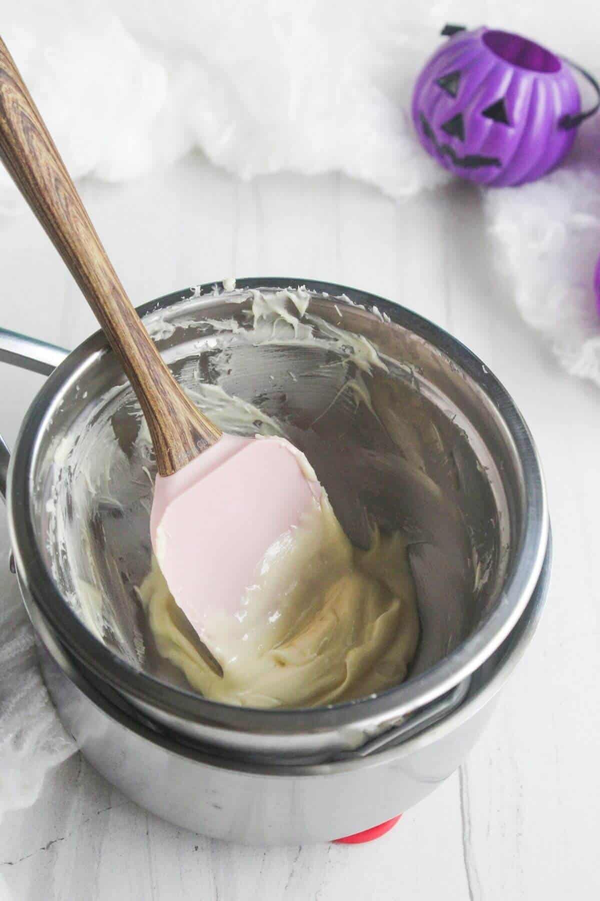 A spatula rests in a metal bowl containing melted white chocolate. Two purple plastic pumpkin containers are in the background.
