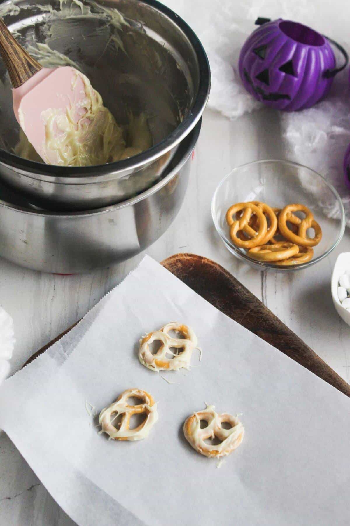 Three pretzels coated in white chocolate on parchment paper, a bowl of plain pretzels, melted chocolate in a double boiler, and a purple pumpkin container on a white surface.