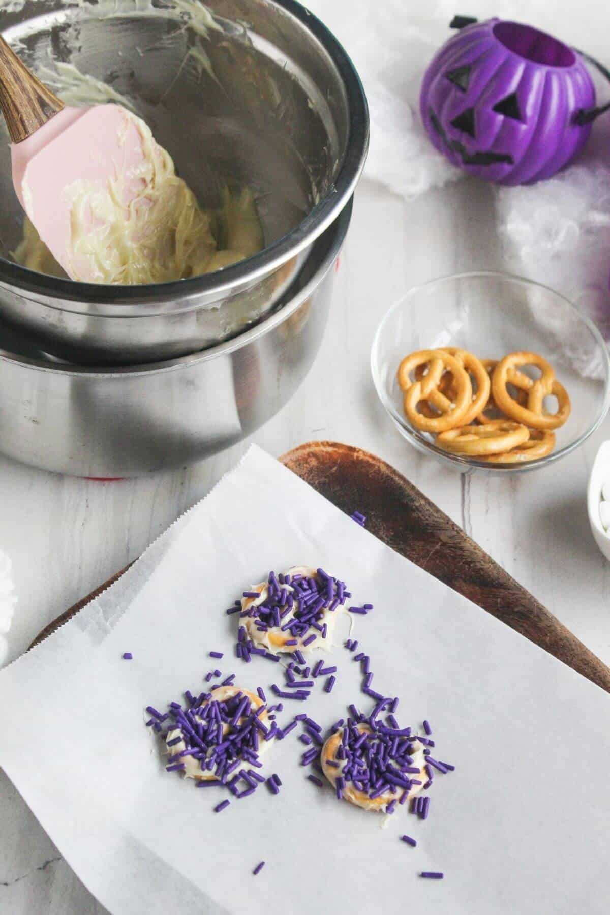 Baking scene with pretzel circles on parchment paper topped with purple sprinkles, a bowl of melted white chocolate, and a purple jack-o'-lantern container in the background.