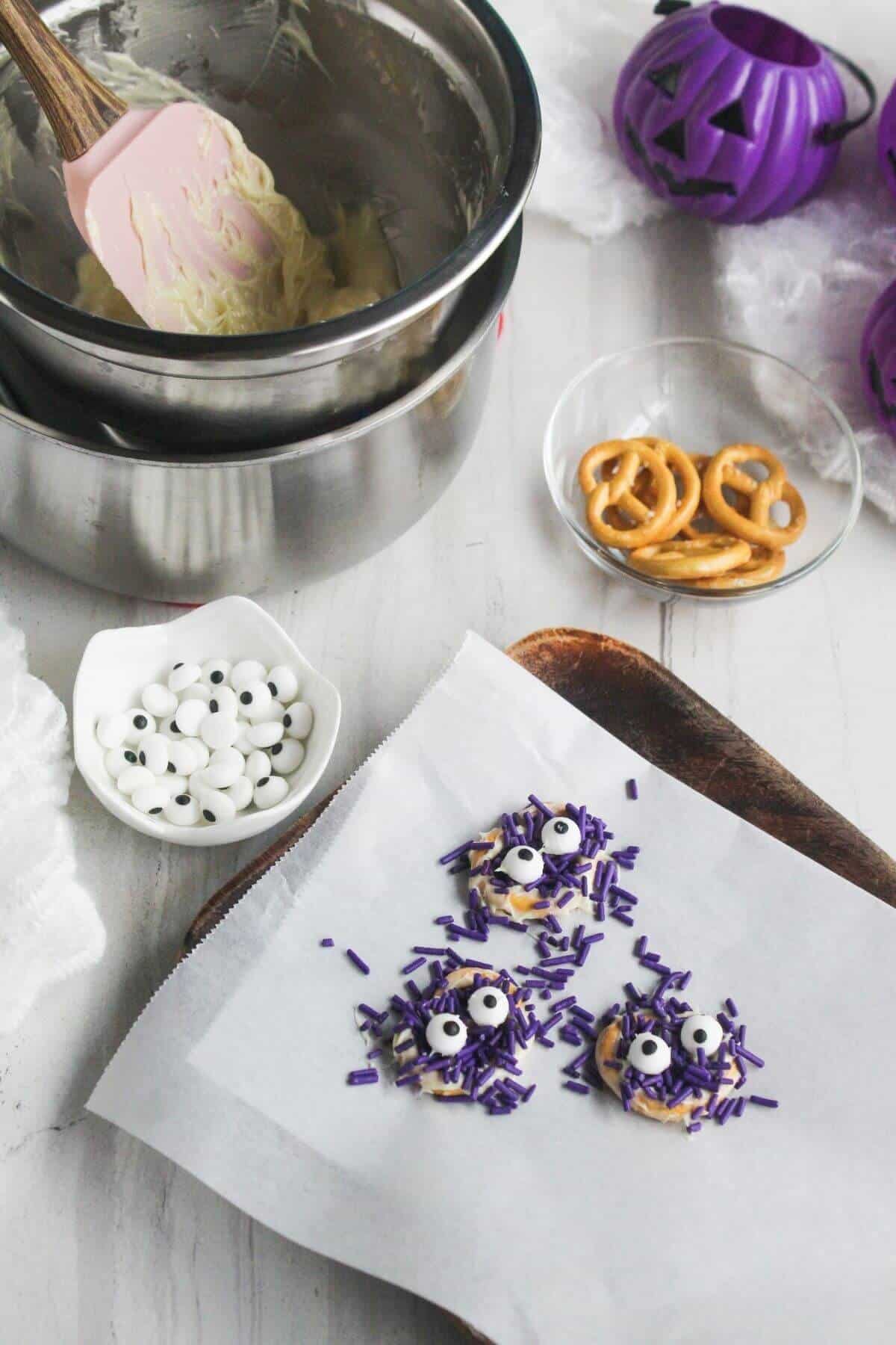 A baking sheet with pretzel treats topped with purple sprinkles and candy eyes. Nearby are bowls with candy eyes, pretzels, melted chocolate, and Halloween decorations.