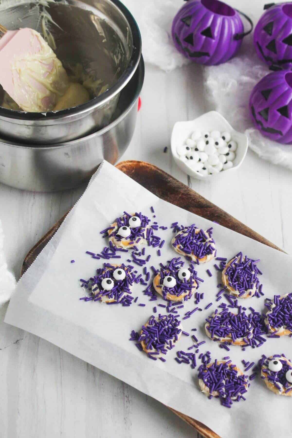 Baking sheet with cookies covered in purple sprinkles and candy eyes, next to a bowl of melted chocolate and purple pumpkin decorations.