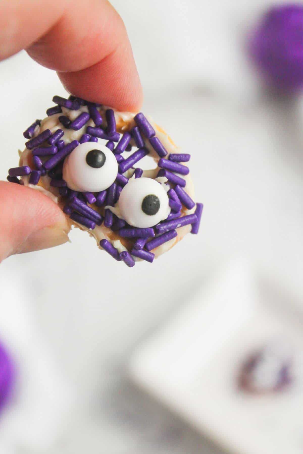A hand holds a round cookie decorated with purple sprinkles and two large candy eyes.