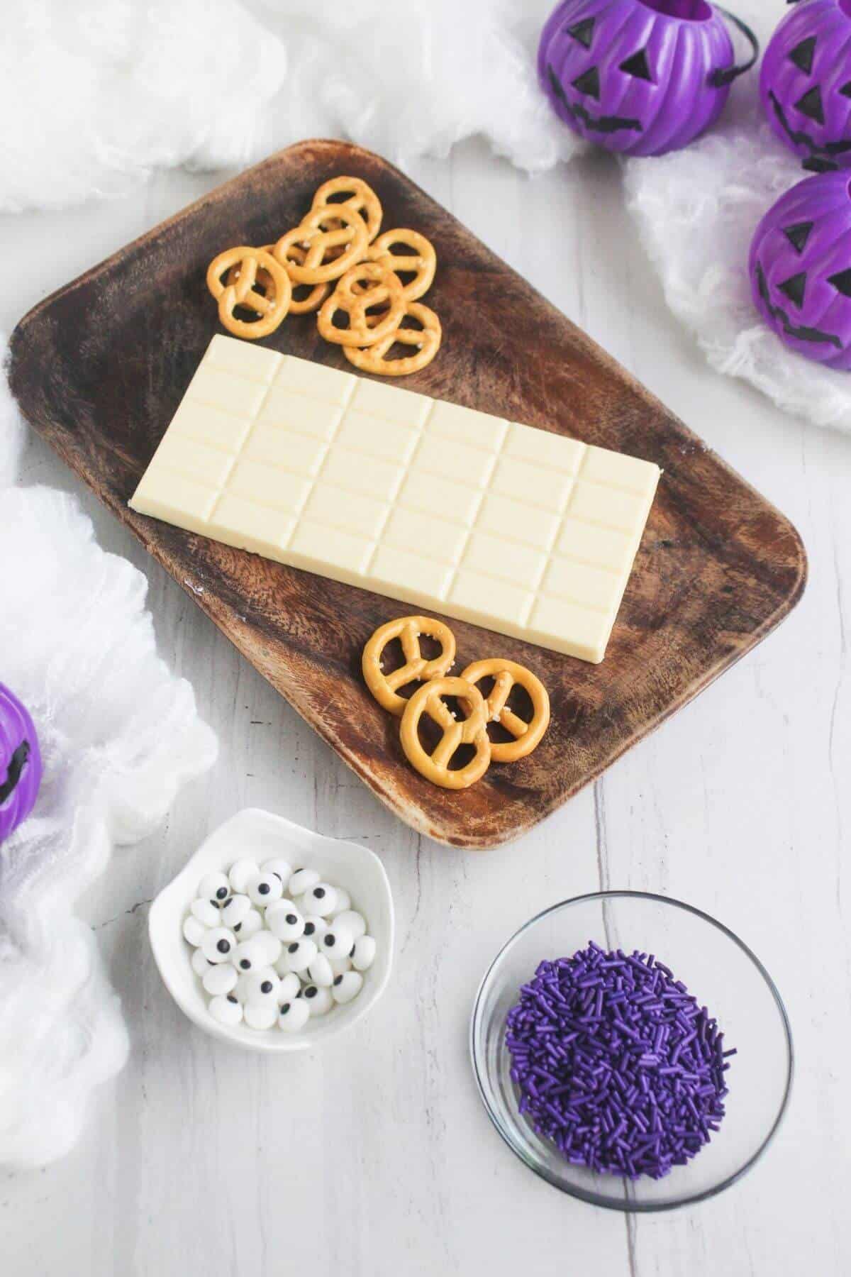 A wooden tray holds a white chocolate bar and pretzels. Nearby are bowls with candy eyes and purple sprinkles, with purple pumpkin decorations and white fabric in the background.