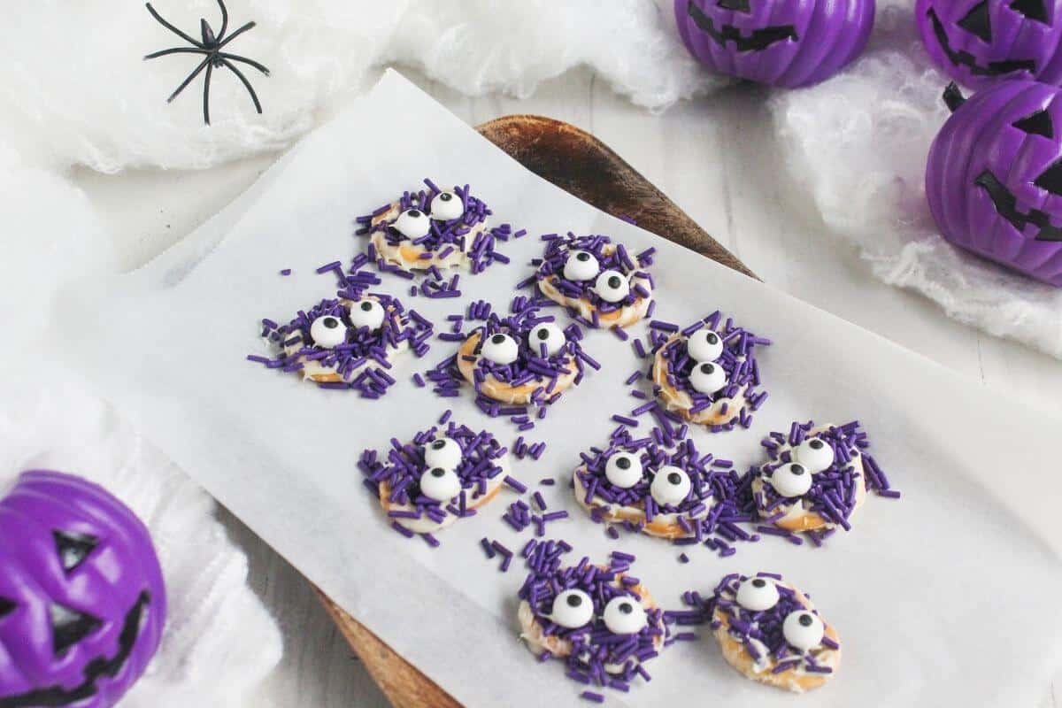 Round cookies with purple sprinkles and candy eyes are arranged on parchment paper. Halloween decorations, including fake spider webs and purple jack-o'-lanterns, are in the background.