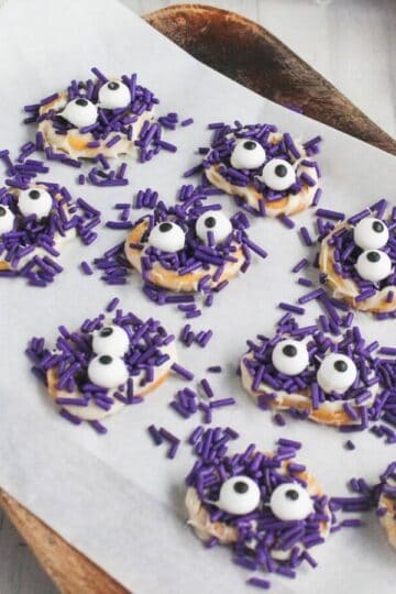 Mini pretzels coated with purple sprinkles and candy eyes are arranged on parchment paper; purple jack-o'-lantern decorations are nearby.