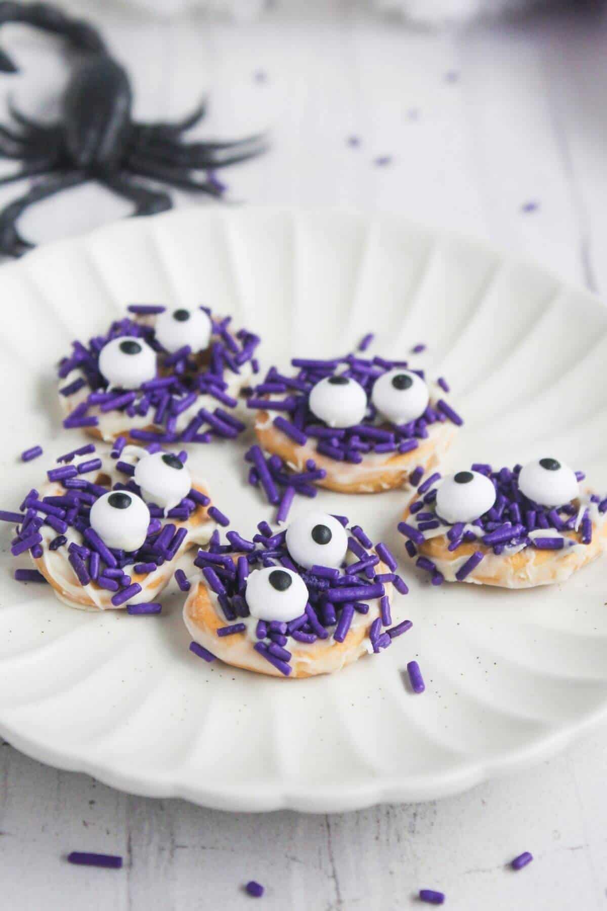 Five small treats with white frosting, purple sprinkles, and candy eyes are arranged on a white plate.