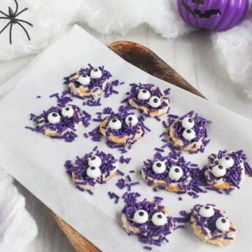 Round cookies decorated with purple sprinkles and candy eyes are arranged on a parchment-lined tray. Halloween-themed decor, including a purple pumpkin, is visible in the background.