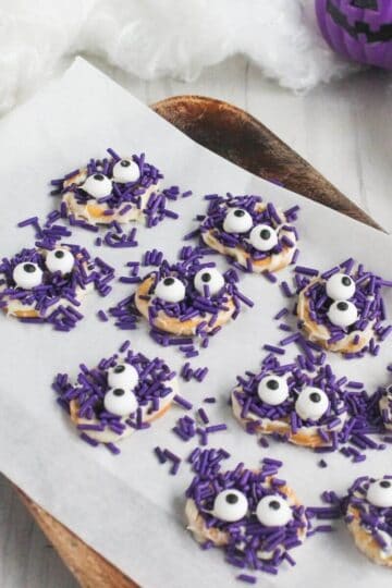 Round cookies decorated with purple sprinkles and candy eyes are arranged on a parchment-lined tray. Halloween-themed decor, including a purple pumpkin, is visible in the background.