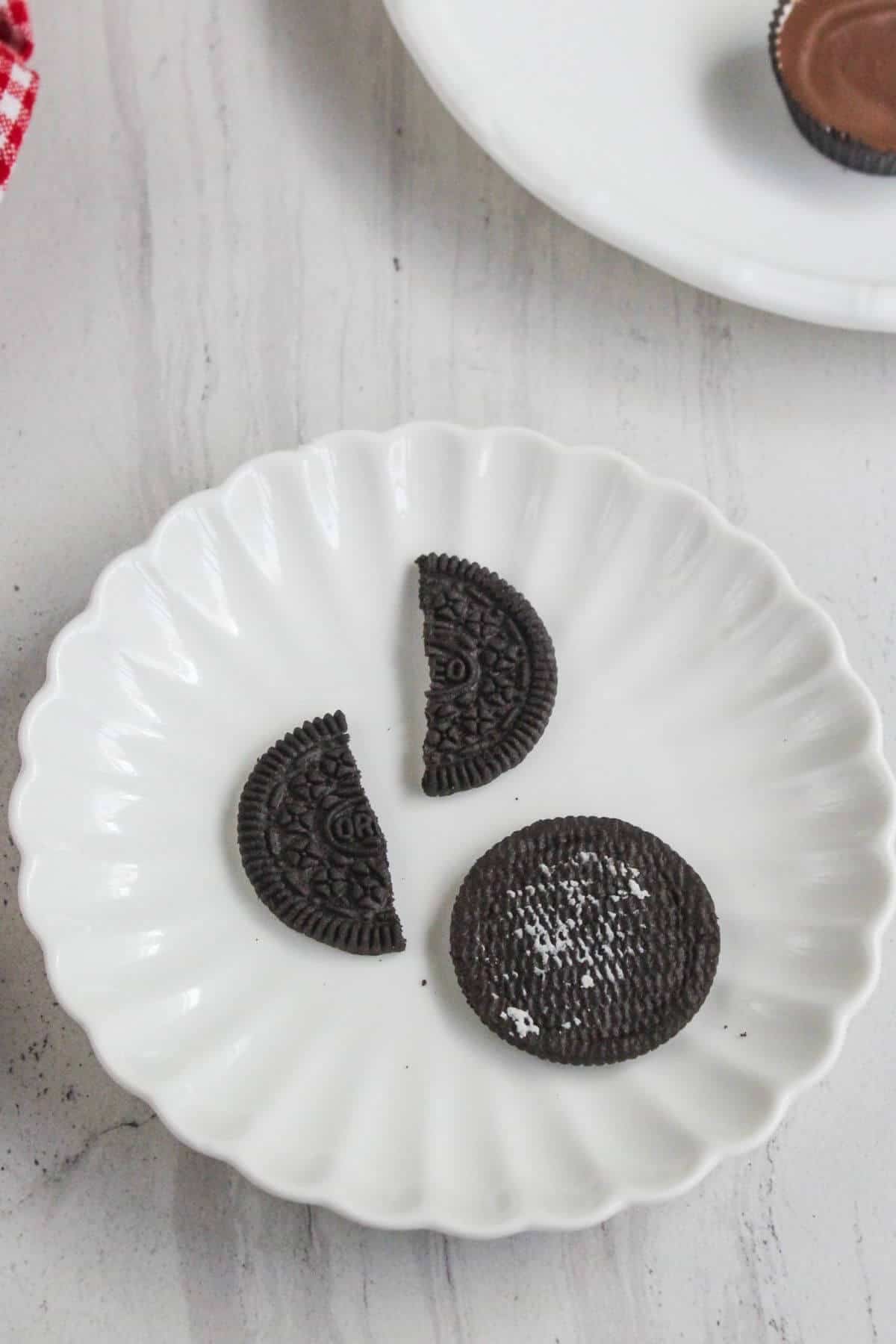 A white plate holds a whole chocolate sandwich cookie, a half cookie, and a quarter cookie piece on a light-colored surface.