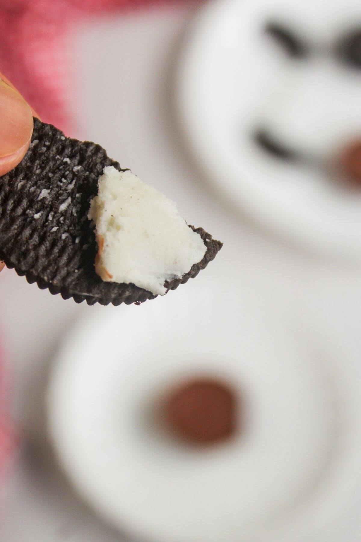 A hand holds a chocolate sandwich cookie half with white cream filling, with blurred plates and cookie pieces in the background.