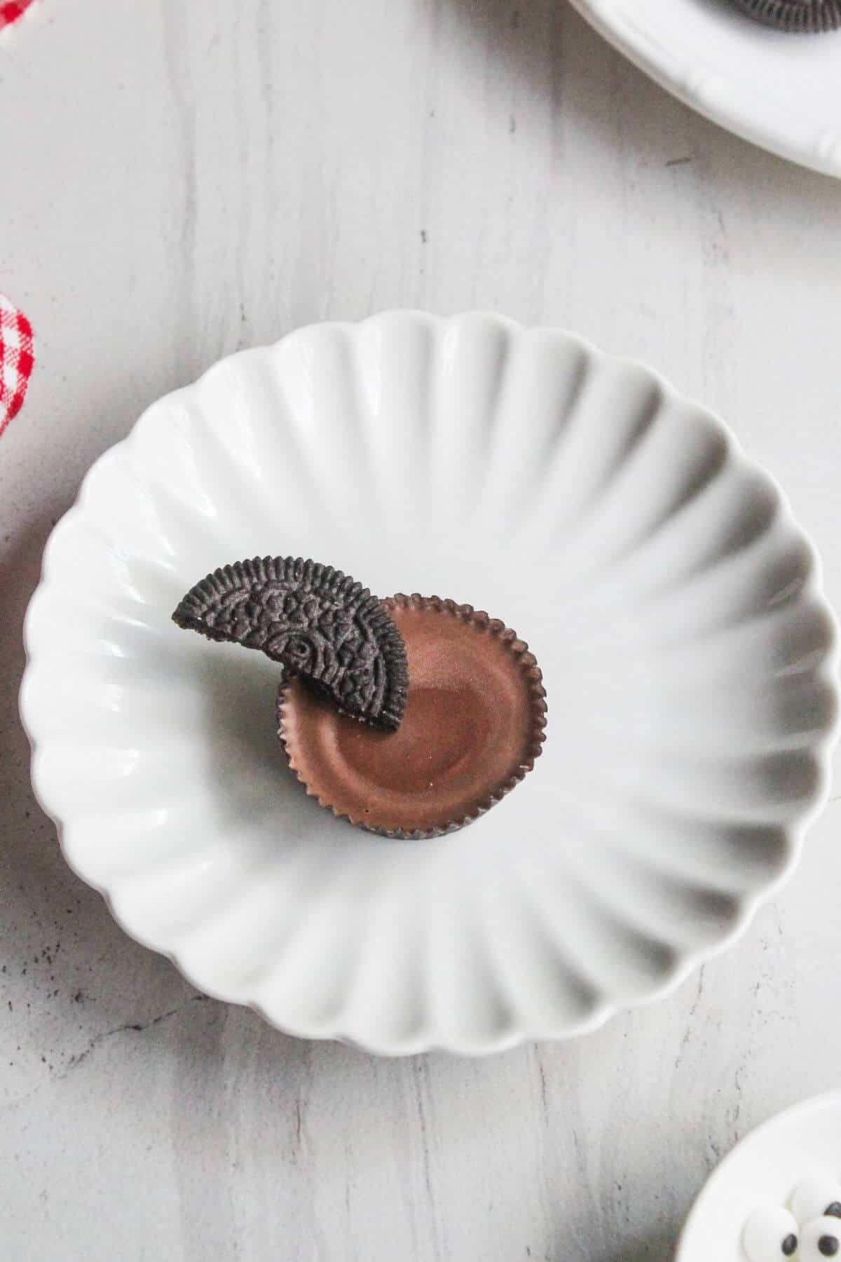 A chocolate peanut butter cup topped with a half piece of chocolate sandwich cookie on a white scalloped plate.