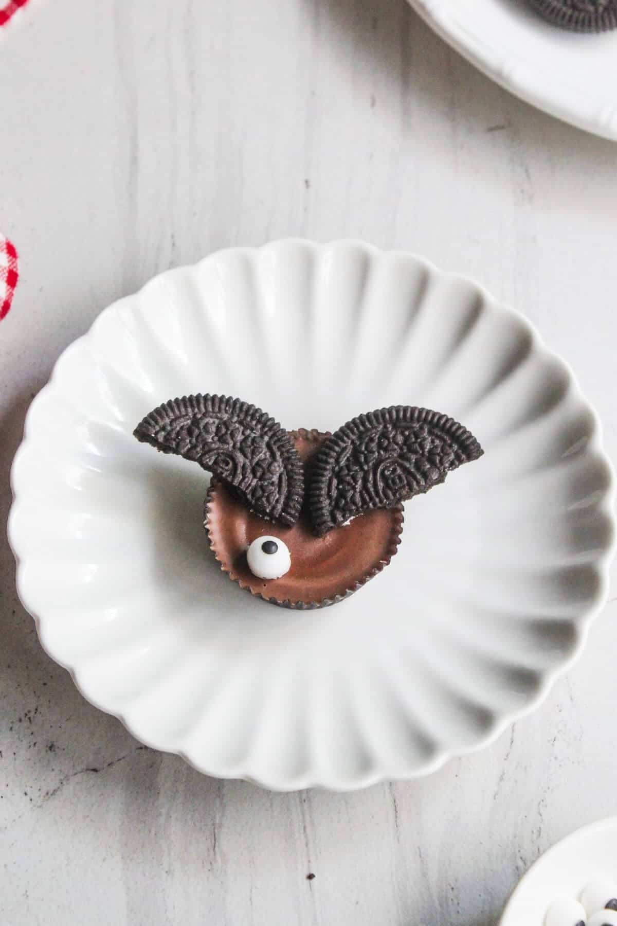 A chocolate cup with two Oreo cookie halves as wings and a candy eye, arranged on a white scalloped plate.
