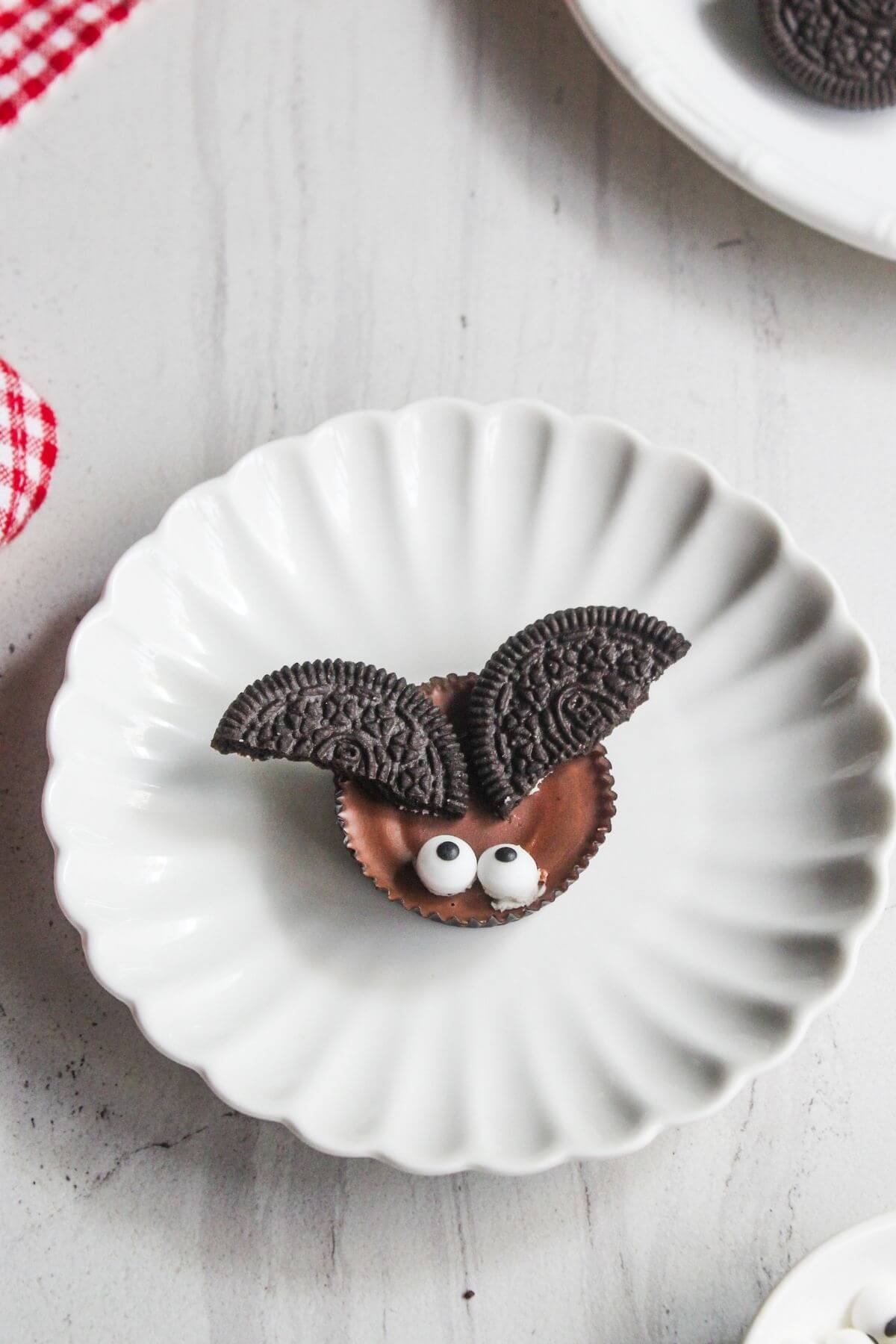 A peanut butter cup with two Oreo halves as ears and two candy eyes arranged to look like a bat on a white plate.