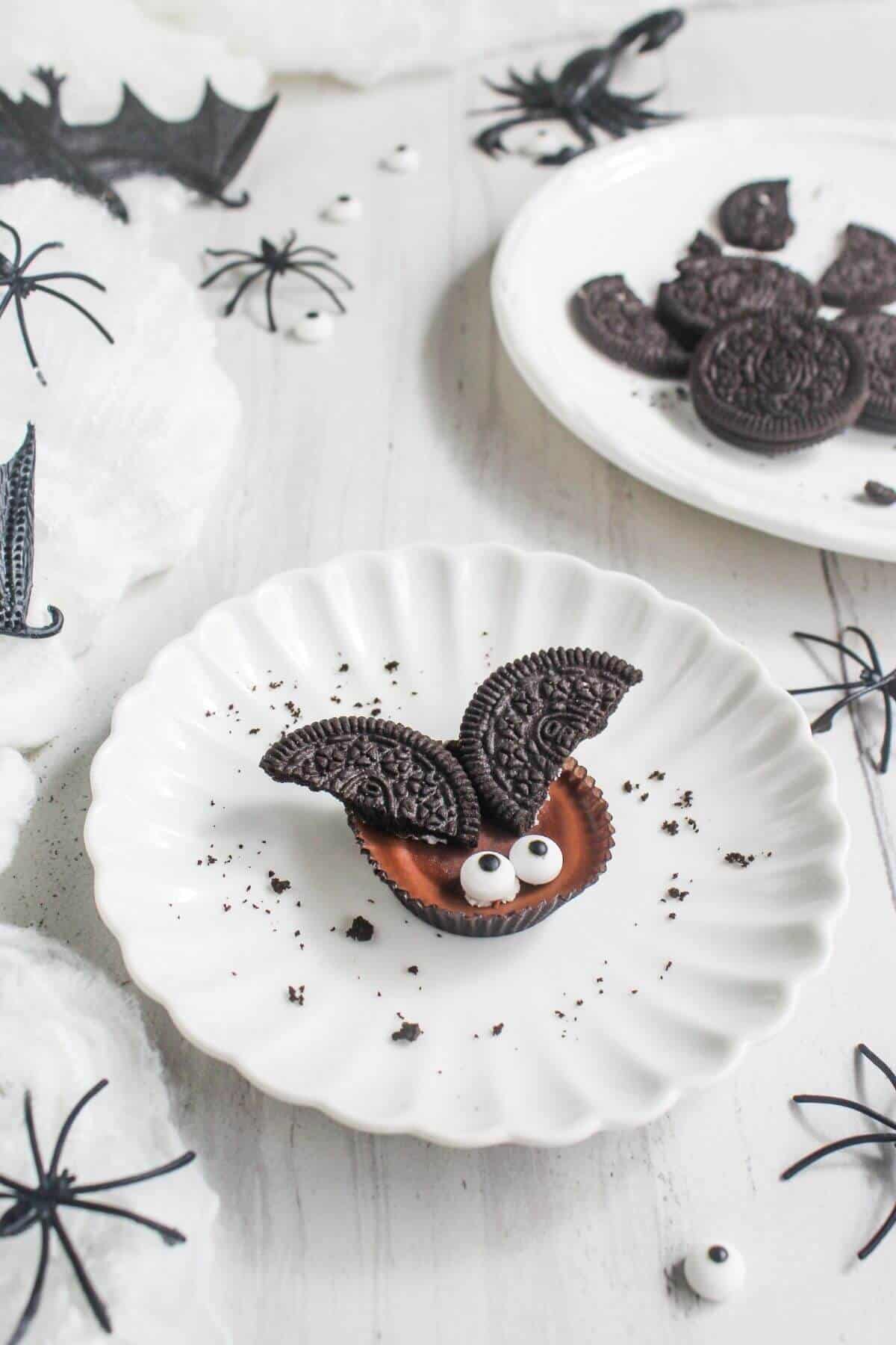 Chocolate cup with two Oreo cookie halves as wings and two candy eyes on a white plate; Halloween decorations and cookies in the background.