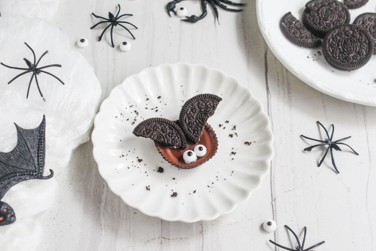 A cookie decorated as a bat with Oreo wings and candy eyes sits on a white plate, surrounded by plastic spiders and more cookies.