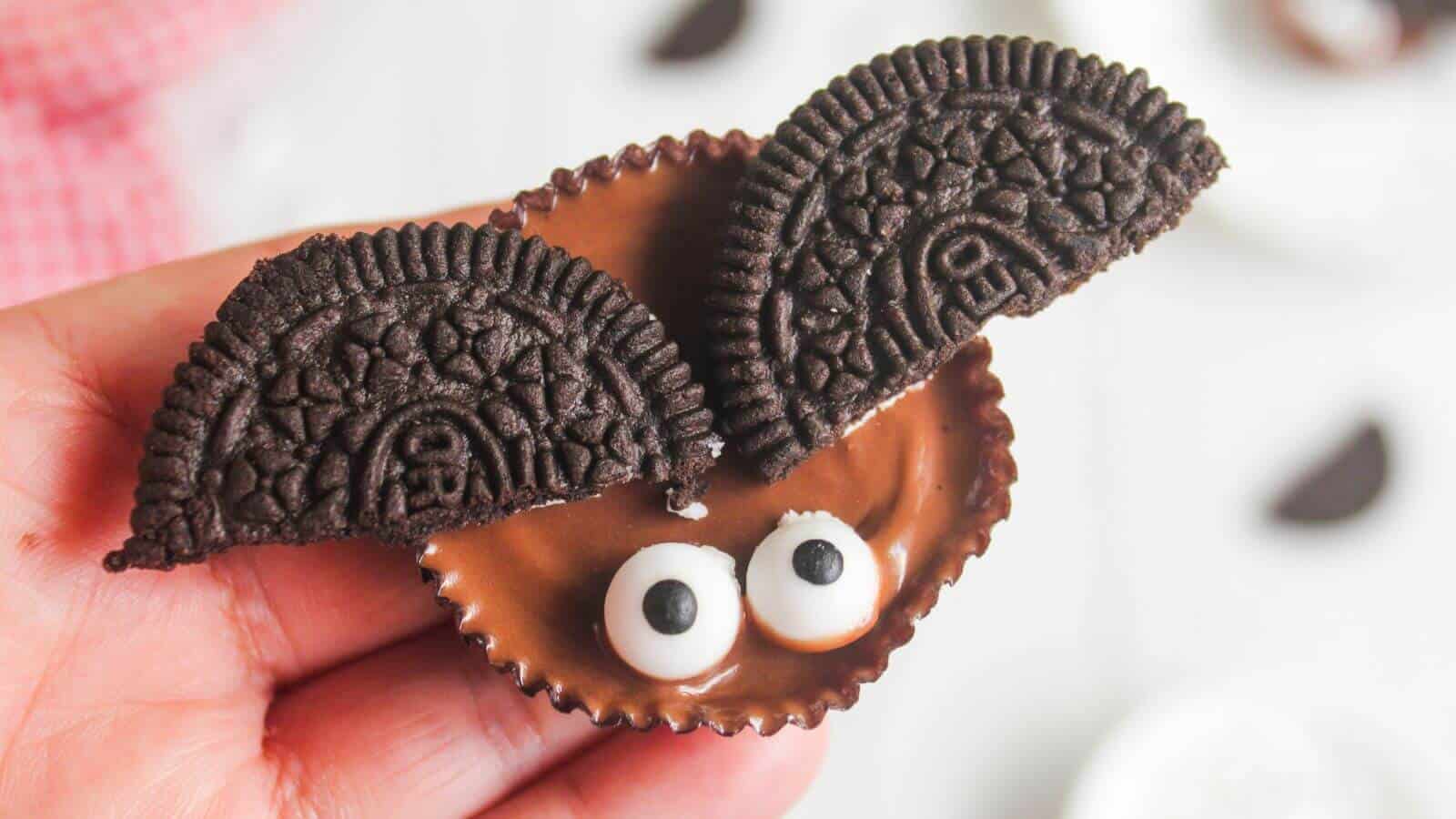 A hand holds a chocolate peanut butter cup decorated with candy eyes and two Oreo cookie halves to resemble a bat.