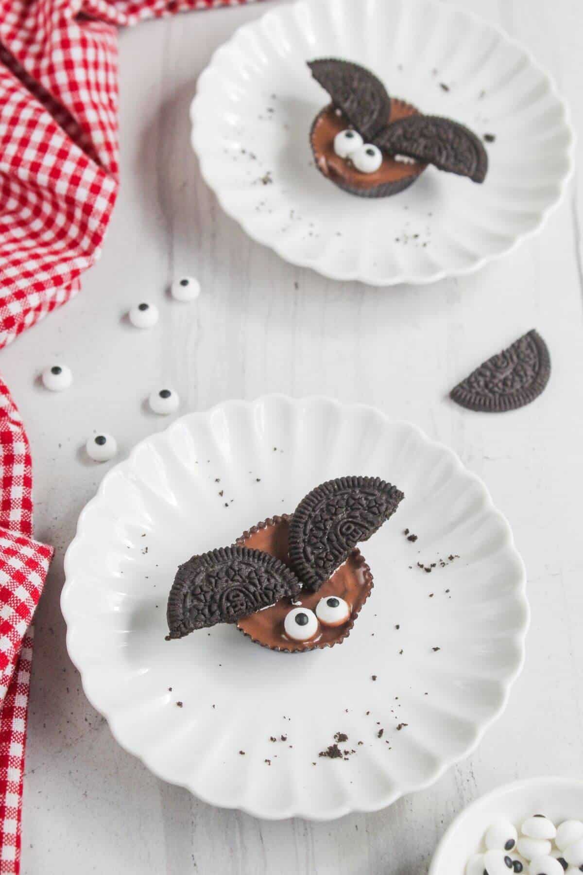 Two white plates each with a chocolate candy decorated with Oreo cookie wings and candy eyes, resembling bats. A red checkered cloth and extra candy eyes are on the table.