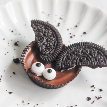 A chocolate candy with two cookie halves as wings and two small candy eyes, arranged to resemble a bat, on a white plate with cookie crumbs.