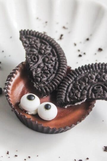 A chocolate candy with two cookie halves as wings and two small candy eyes, arranged to resemble a bat, on a white plate with cookie crumbs.