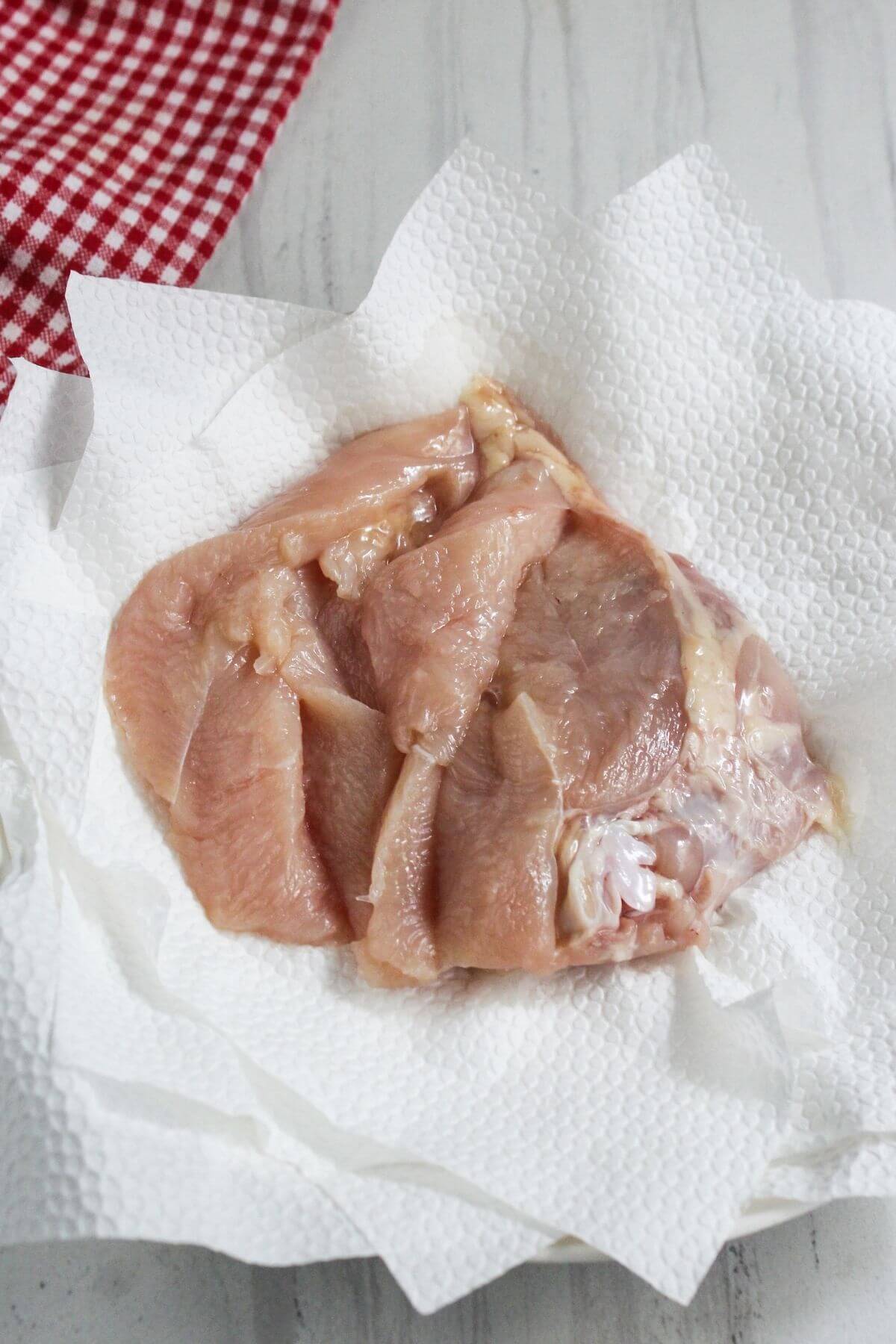Raw chicken breast pieces on a sheet of white paper towel, with a red and white checkered cloth partially visible in the top left corner.