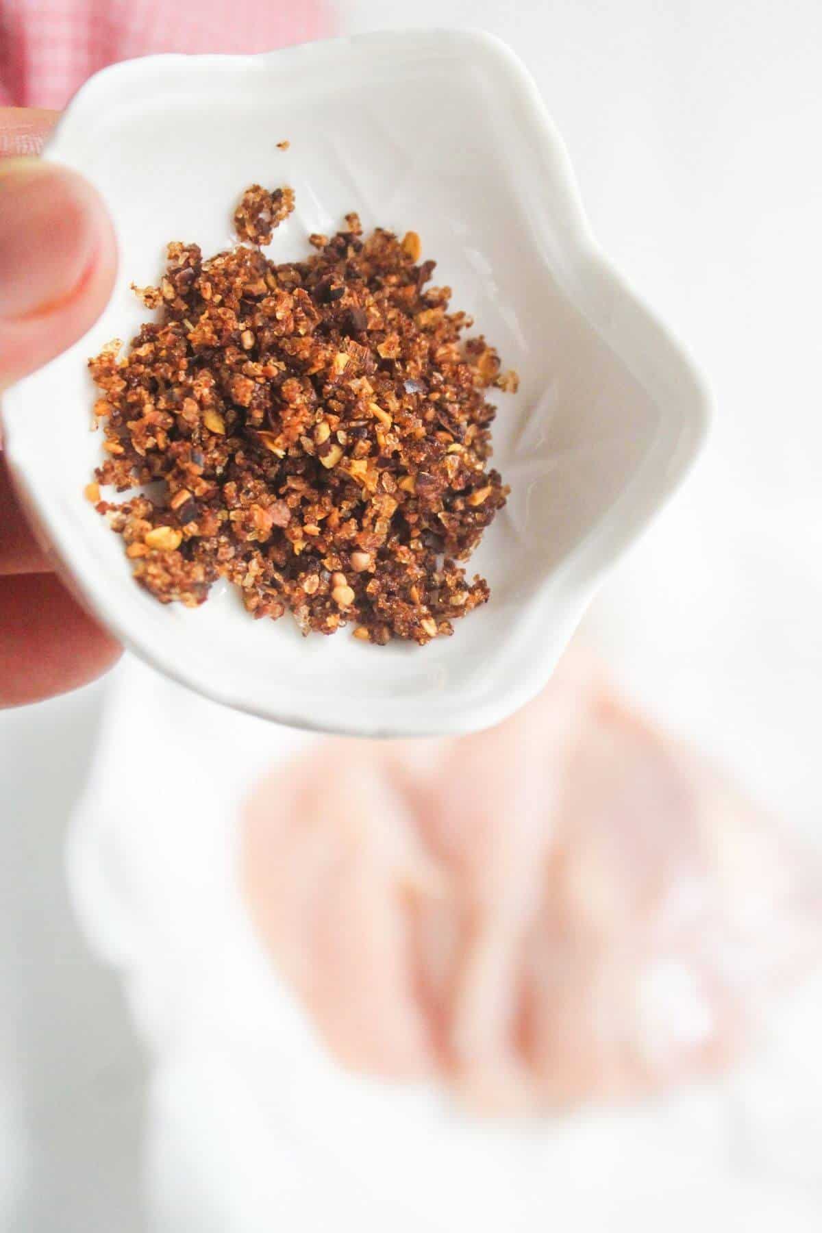 A small white dish holds a coarse, reddish-brown spice blend above raw chicken breasts on a plate in the background.
