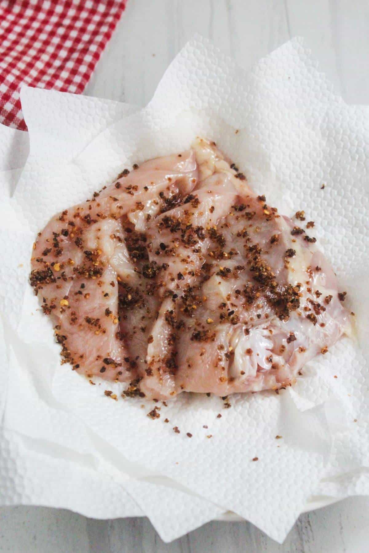 Raw chicken pieces coated with spices rest on white paper towels, with a red and white checkered cloth partially visible in the corner.