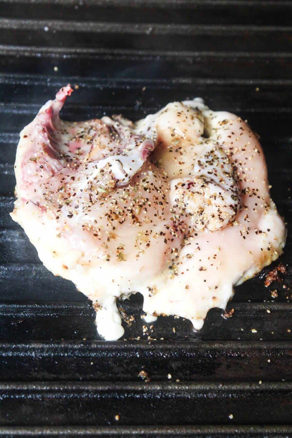 Raw chicken seasoned with black pepper and herbs cooking on a grill, with some juices and seasoning visible on the surface.