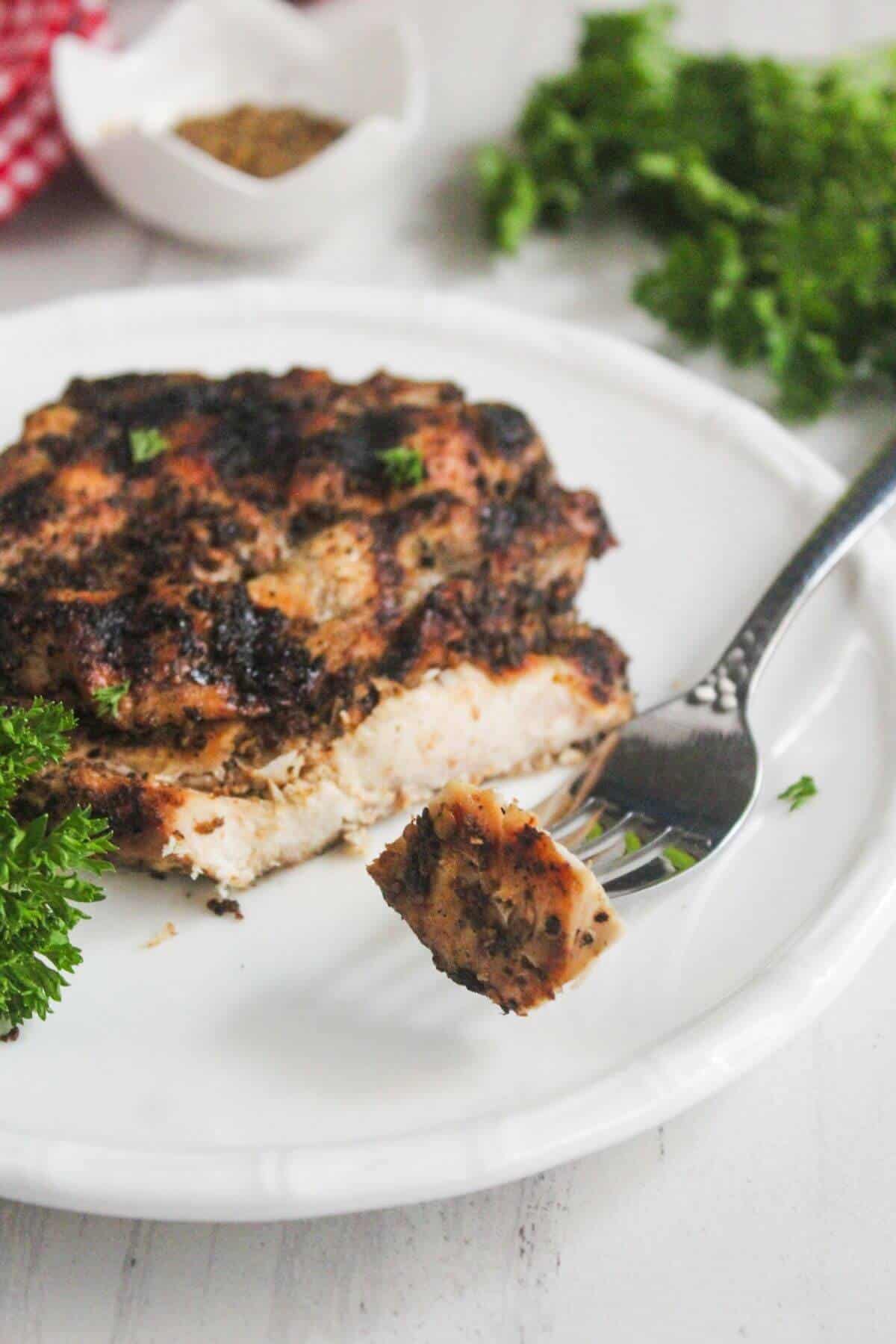 A piece of seasoned, grilled chicken breast on a white plate with a fork holding a bite, garnished with fresh parsley.