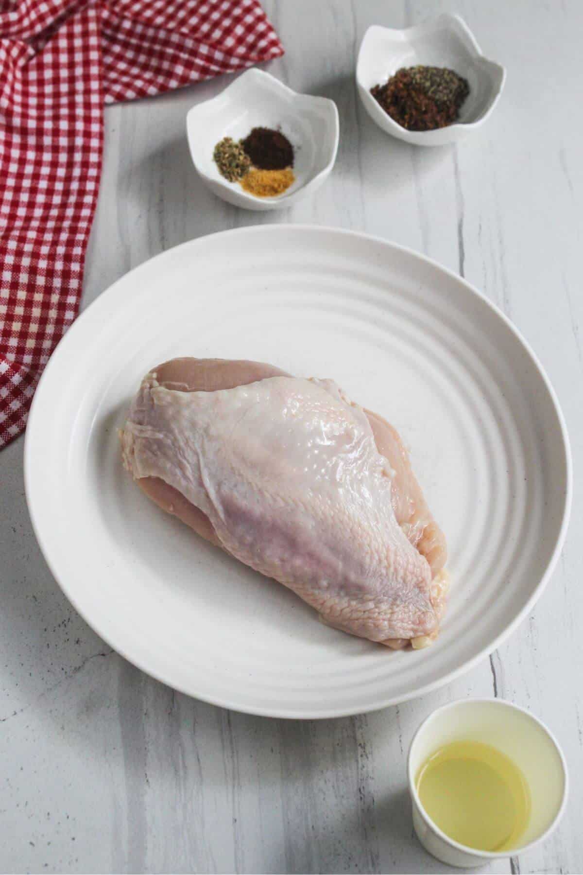 Raw chicken breast on a white plate, with a cup of oil and two small bowls of spices beside a red checkered cloth on a white surface.