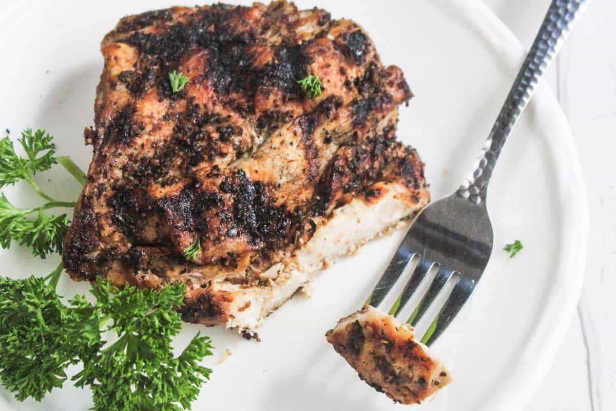 Grilled seasoned chicken breast on a white plate with a fork holding a cut piece, garnished with fresh parsley.