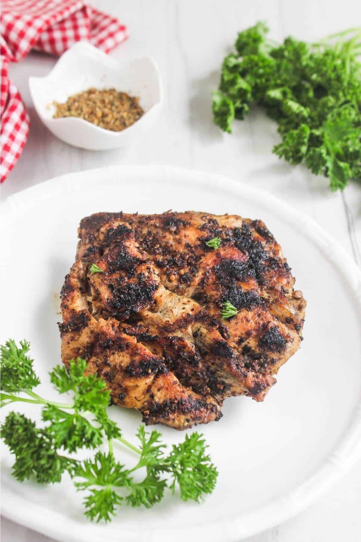 A grilled chicken thigh garnished with parsley on a white plate, with fresh parsley and a bowl of spices in the background.