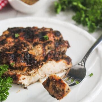 Grilled, seasoned chicken breast on a white plate with a piece cut and held by a fork; parsley garnish is visible nearby.