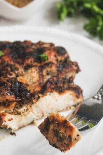 Grilled, seasoned chicken breast on a white plate with a piece cut and held by a fork; parsley garnish is visible nearby.
