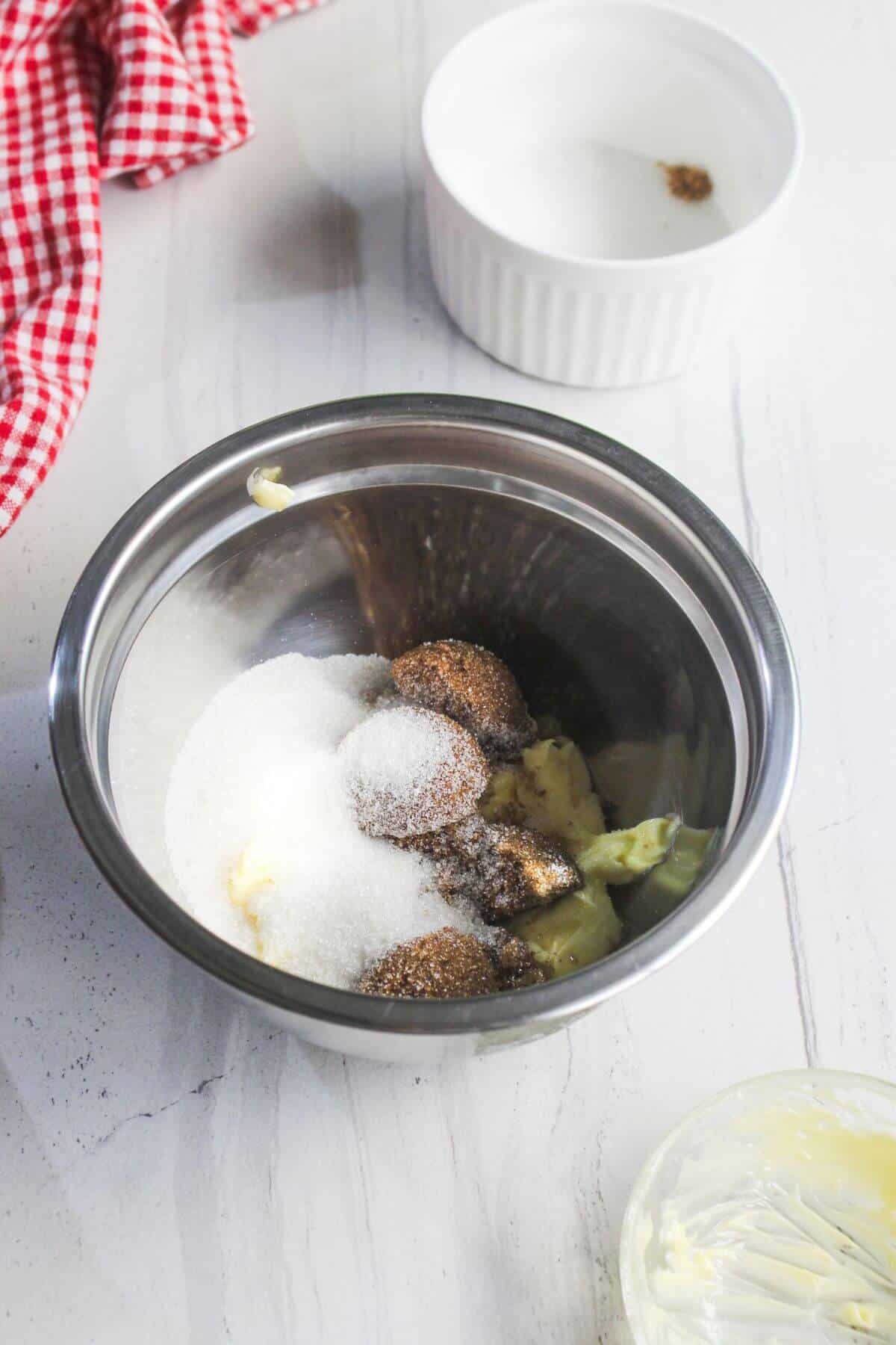 A metal bowl with butter, white sugar, and brown sugar sits on a white countertop next to a glass dish, a ramekin with spices, and a red checkered cloth.