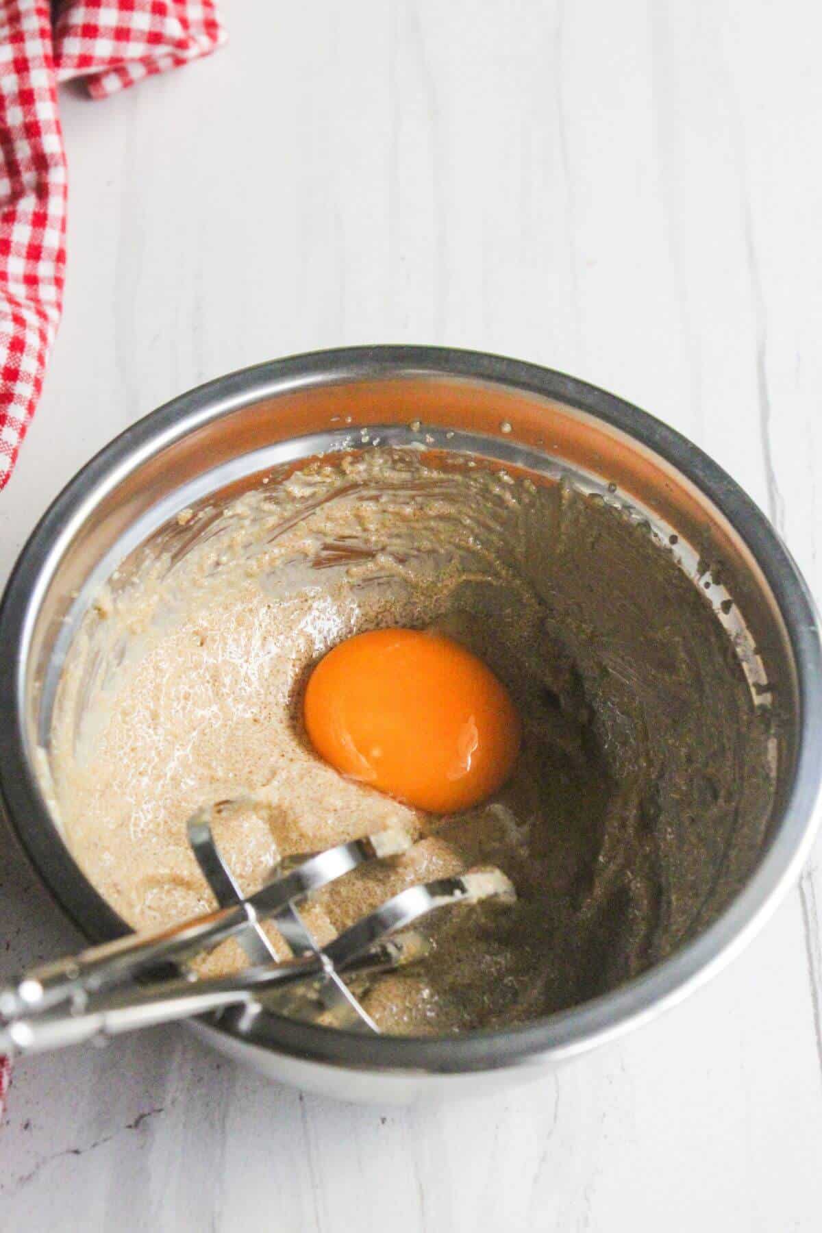 A metal mixing bowl with batter, a raw egg yolk, and a hand mixer on a white surface. A red and white cloth is nearby.