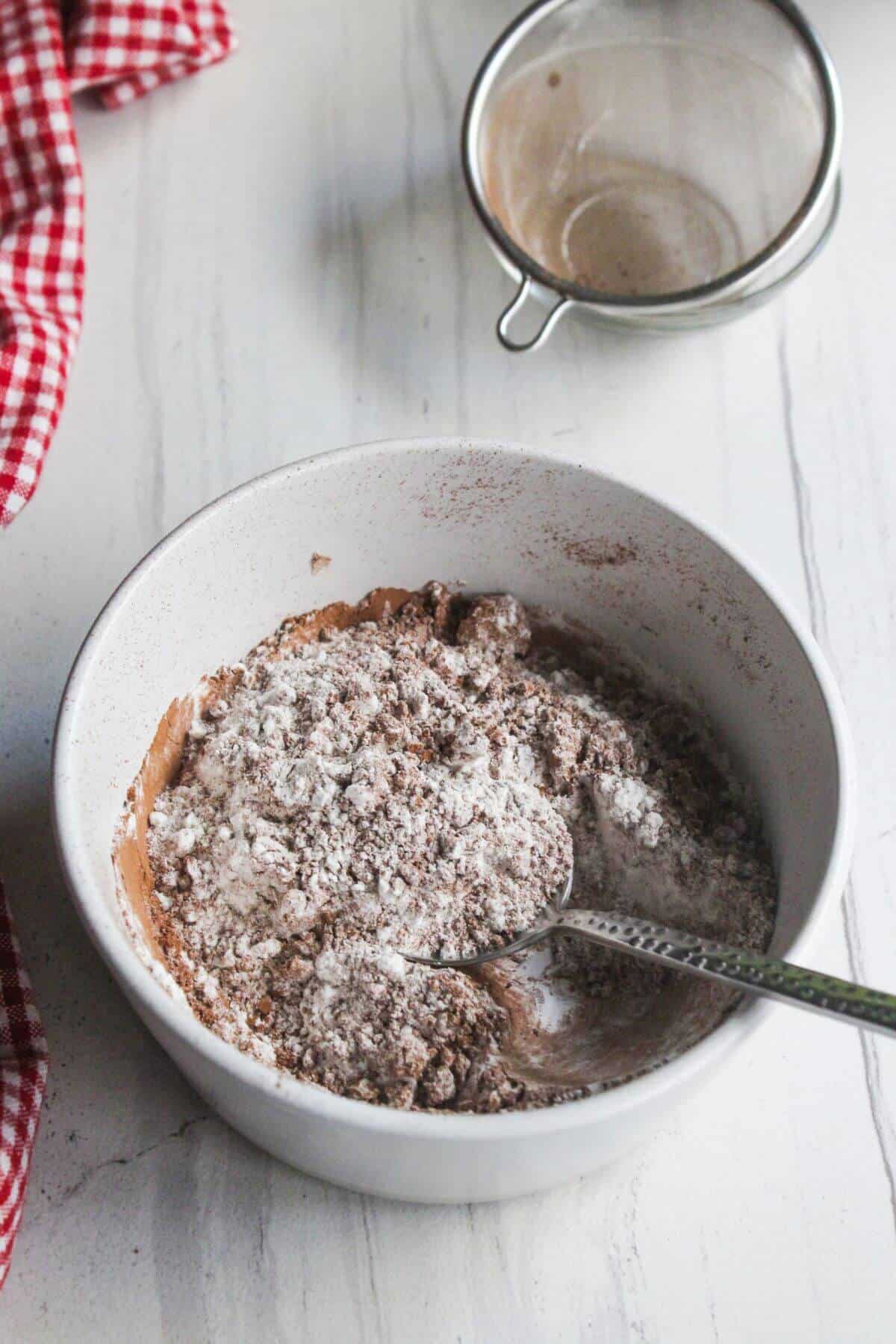 A white bowl with flour and cocoa powder being mixed with a spoon sits on a white countertop; a mesh sifter and red checkered cloth are nearby.
