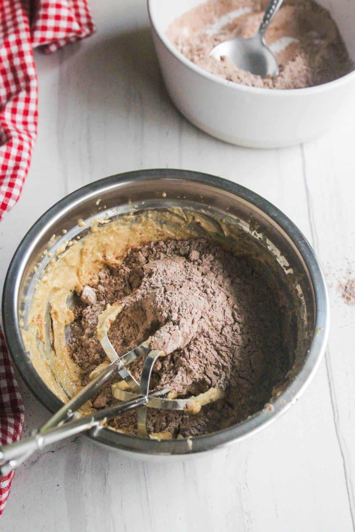 A metal bowl with partially mixed chocolate cake batter and a hand mixer sits on a white surface; a bowl of dry ingredients and a red-checkered cloth are nearby.