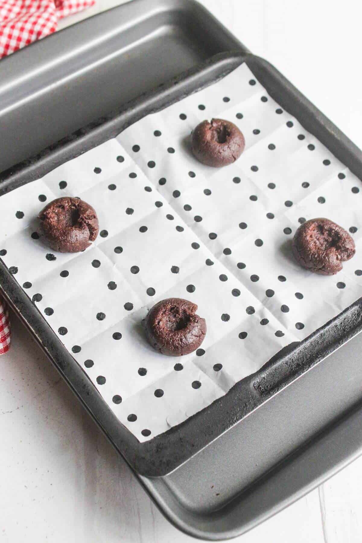 A baking tray lined with polka dot parchment paper holds four unbaked chocolate cookie dough balls spaced apart.