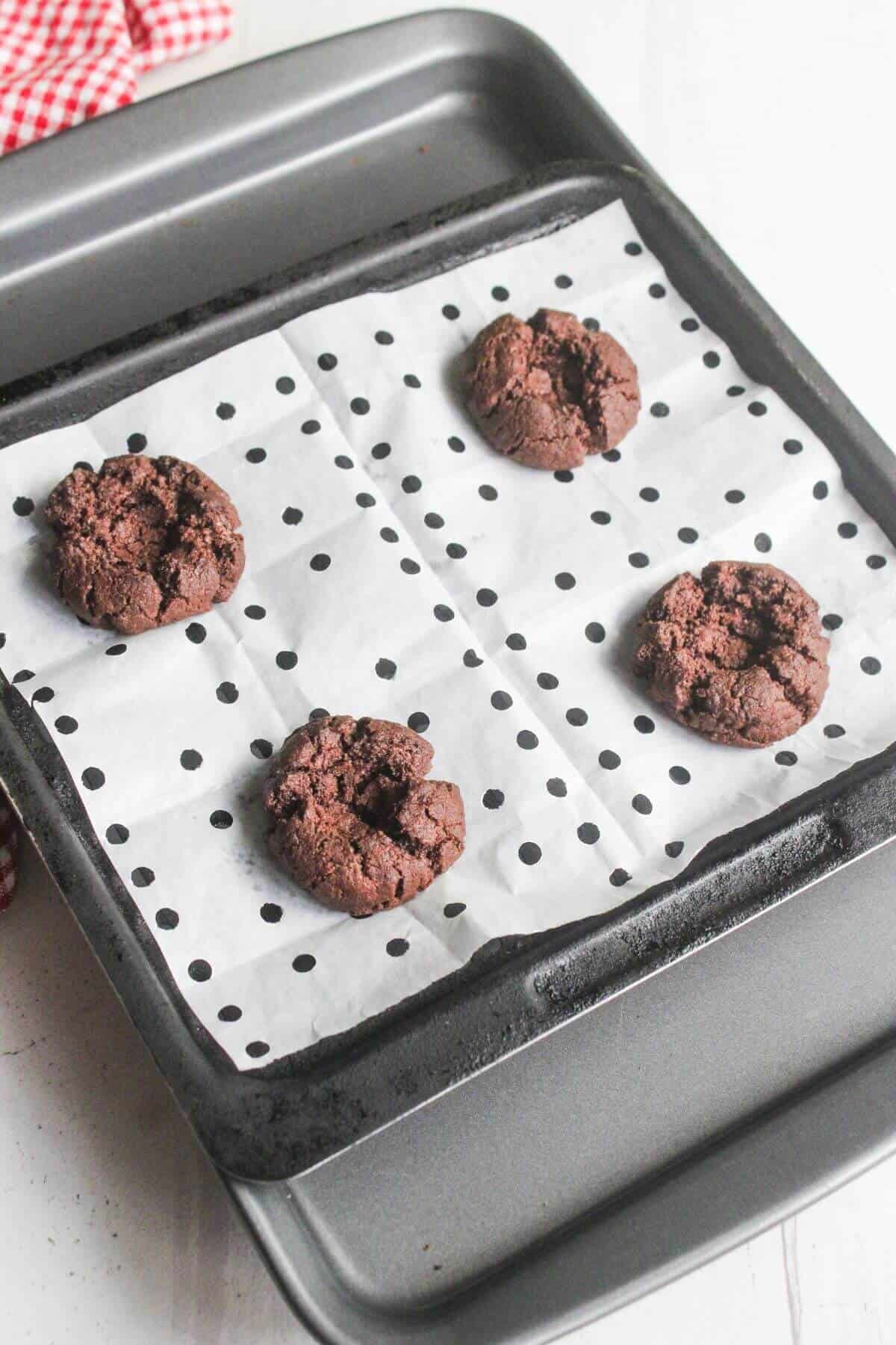 A baking tray lined with dotted parchment paper holds four chocolate cookies spaced apart.
