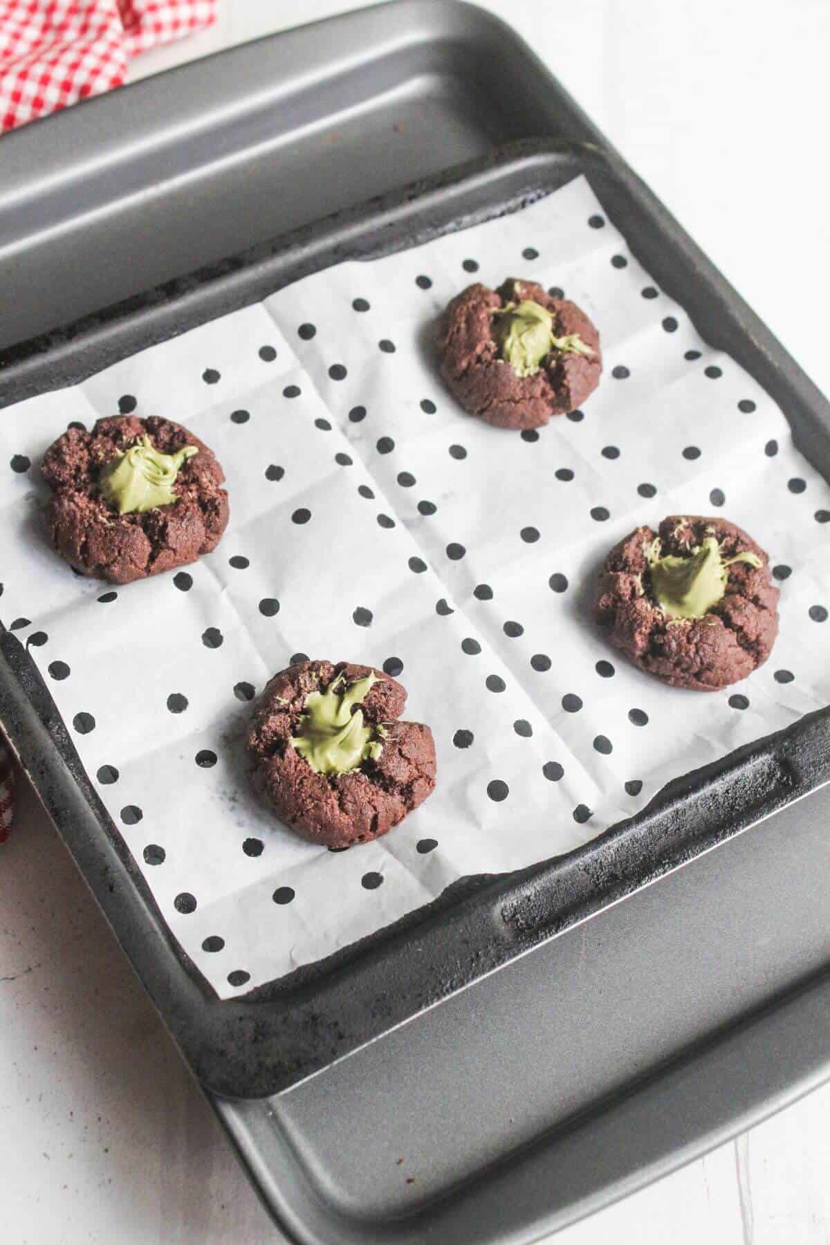 A baking tray with four chocolate cookies topped with green filling sits on polka dot parchment paper.