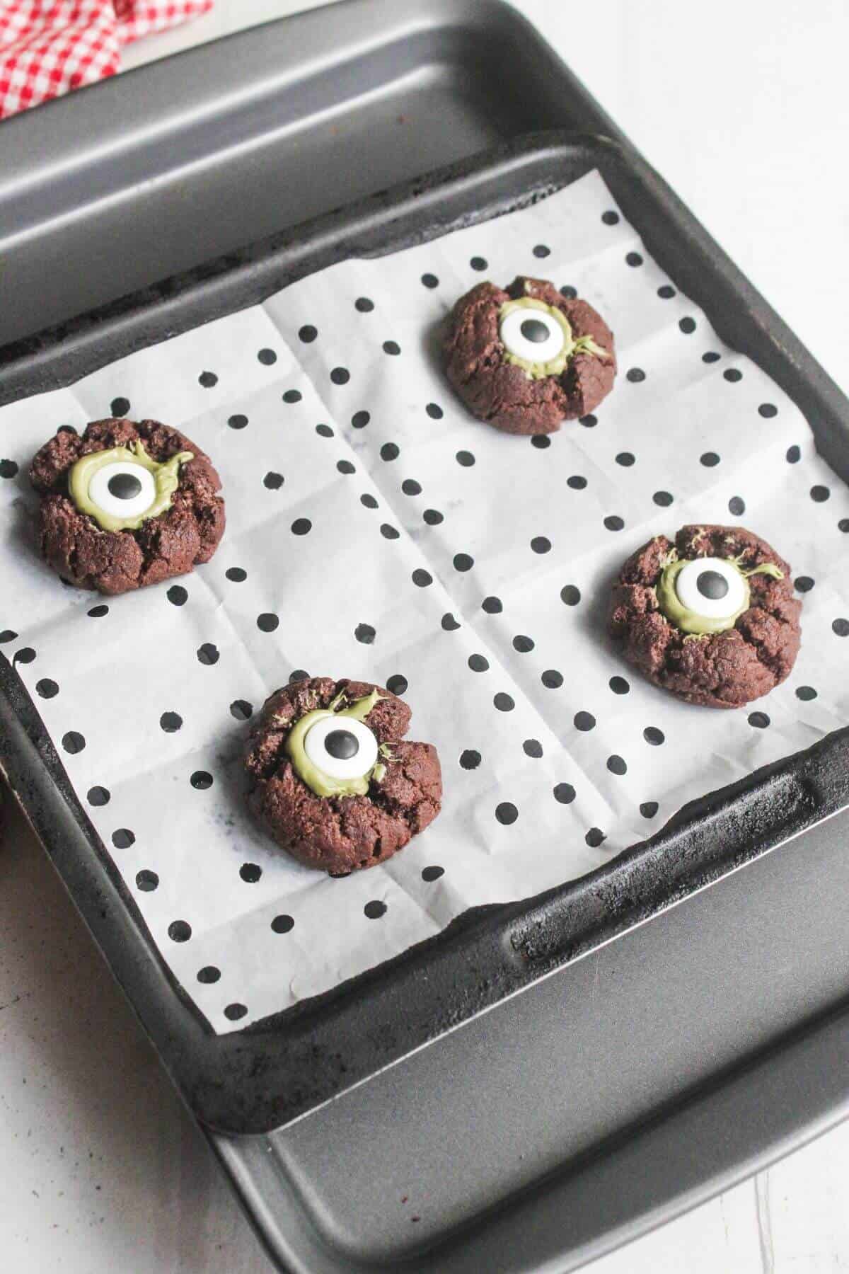 A baking tray with four chocolate cookies topped with green and white candy eyes, placed on polka dot parchment paper.