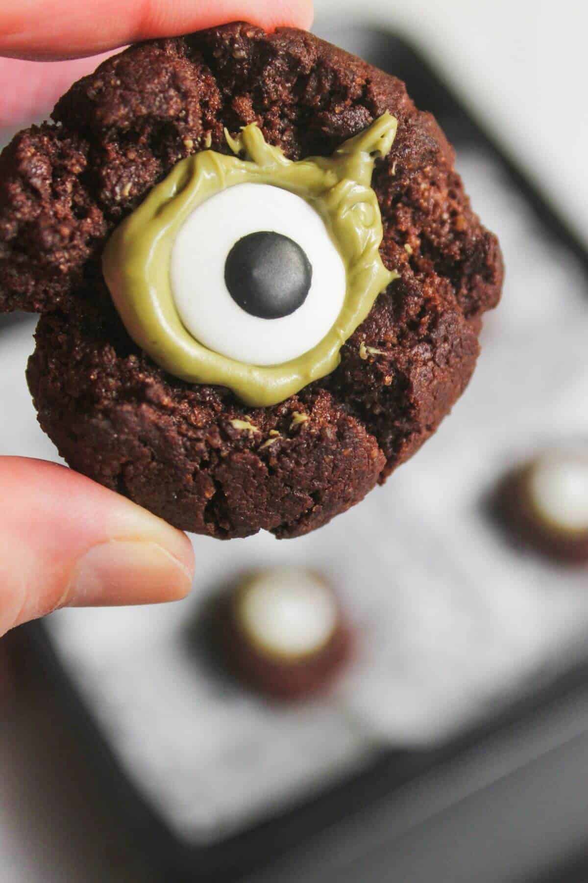 A hand holds a chocolate cookie decorated with green icing and a candy eye to resemble a monster eye. A baking tray with more cookies is blurred in the background.