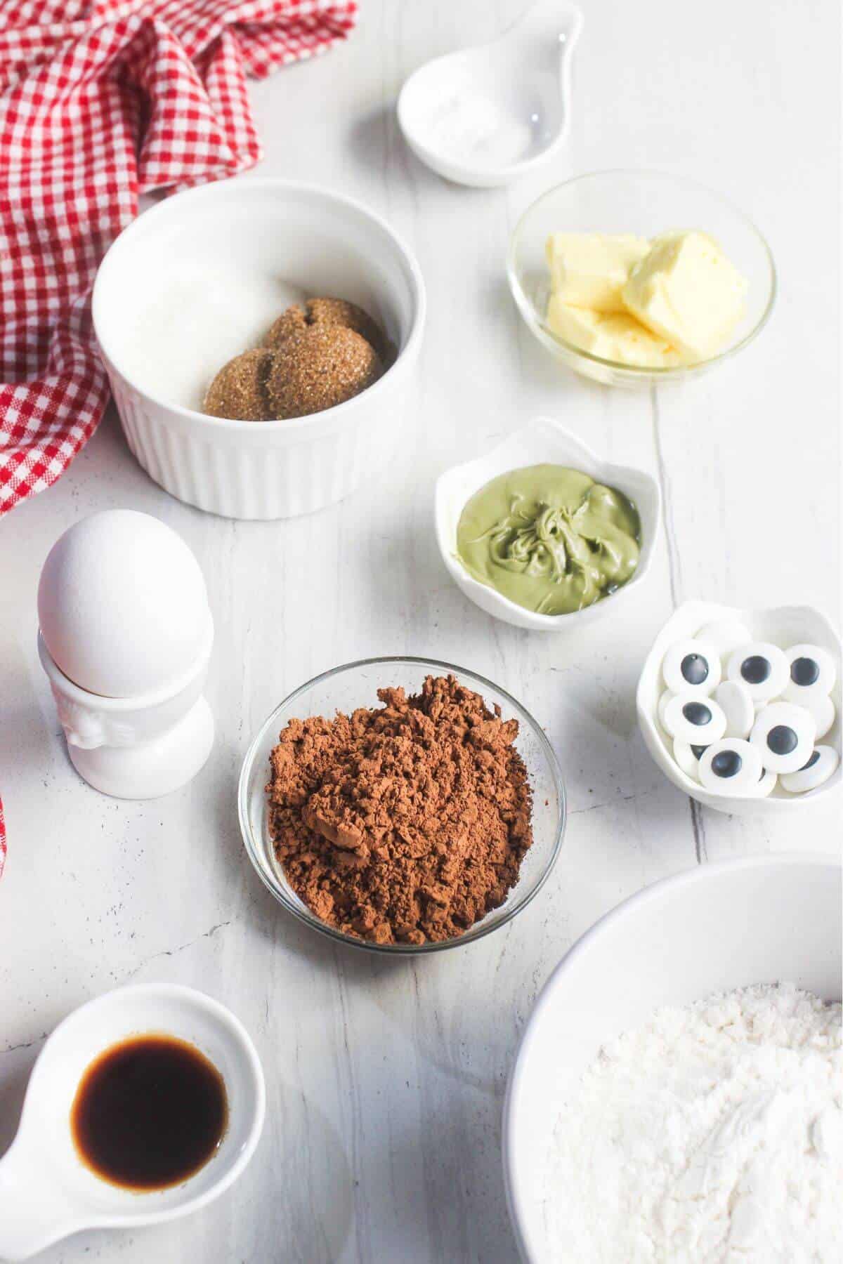 Various baking ingredients in small bowls, including flour, cocoa powder, sugars, butter, vanilla, an egg, green cream, candy eyes, and a red checkered cloth on a white surface.